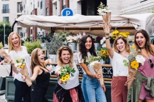 Dzień Kobiet w pracy. Zdjęcie przedstawia 6 radosnych kobiet trzymających żółte kwiaty. W tle spokojna uliczka, stragan z kwiatami.